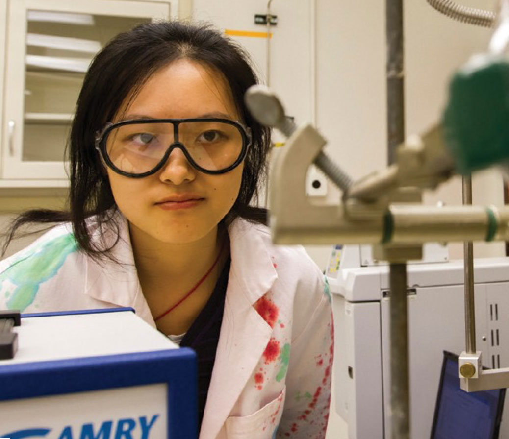Young undergraduate woman sits in a lab in her lab coat and safety goggles looking at a piece of lab equipment