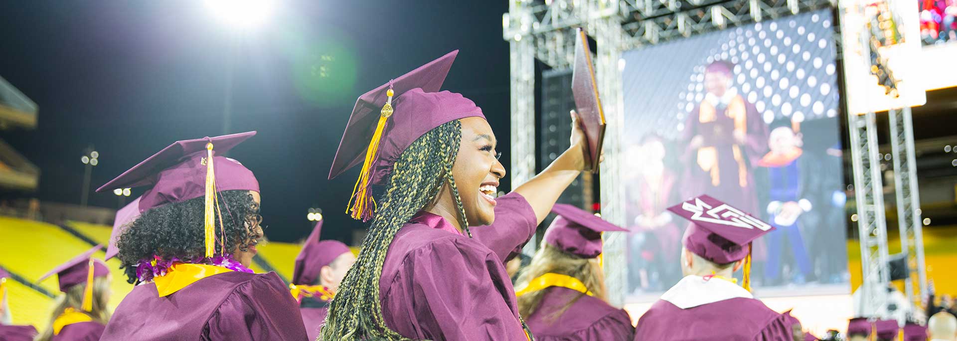 Black female student seated during Convocation and waving at someone while smiling.