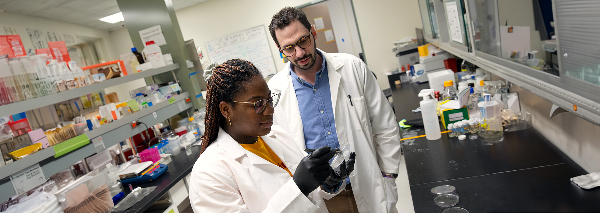 Black female student in a lab coat talking to her male professor in a lab setting.