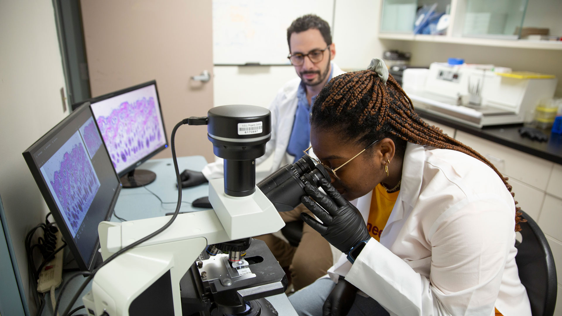 A woman in a lab environment looks into a microscope while her advisor sits next to her. Two screens display images, presumably from the microscope.